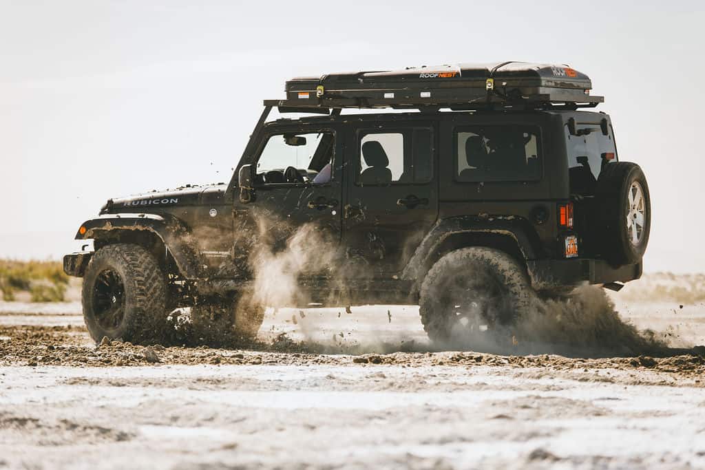 Black Jeep Wrangler JK Rubicon driving through wet sand with a Front Runner Slimline II roof rack and RoofNest rooftop tent on top, kicking up spray.