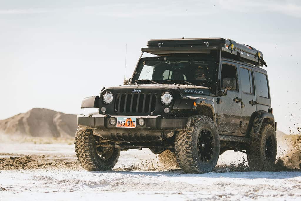 Front three-quarter view of a black Jeep Wrangler JK Rubicon driving fast through wet sand with a Front Runner Slimline II rack and rooftop tent on top.
