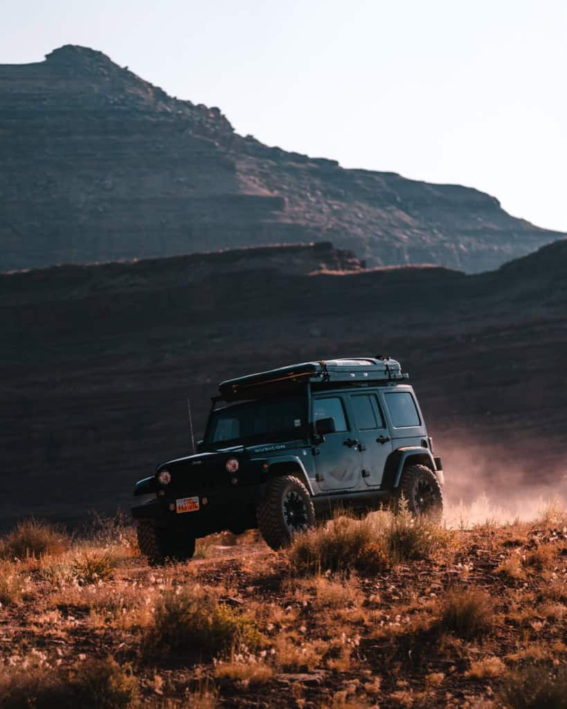 Black Jeep Wrangler JK Rubicon driving on a Moab desert ridge with a hard-shell rooftop tent on the Front Runner roof rack, kicking up dust, backlit by golden side-light.